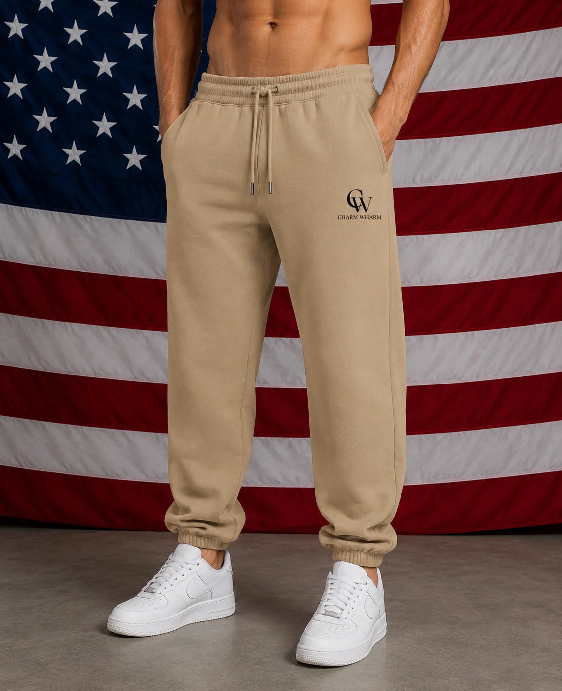 A person wearing beige jogger sweatpants with a drawstring waist and a small black 'CW' logo on the left thigh, paired with white sneakers, standing in front of an American flag backdrop.