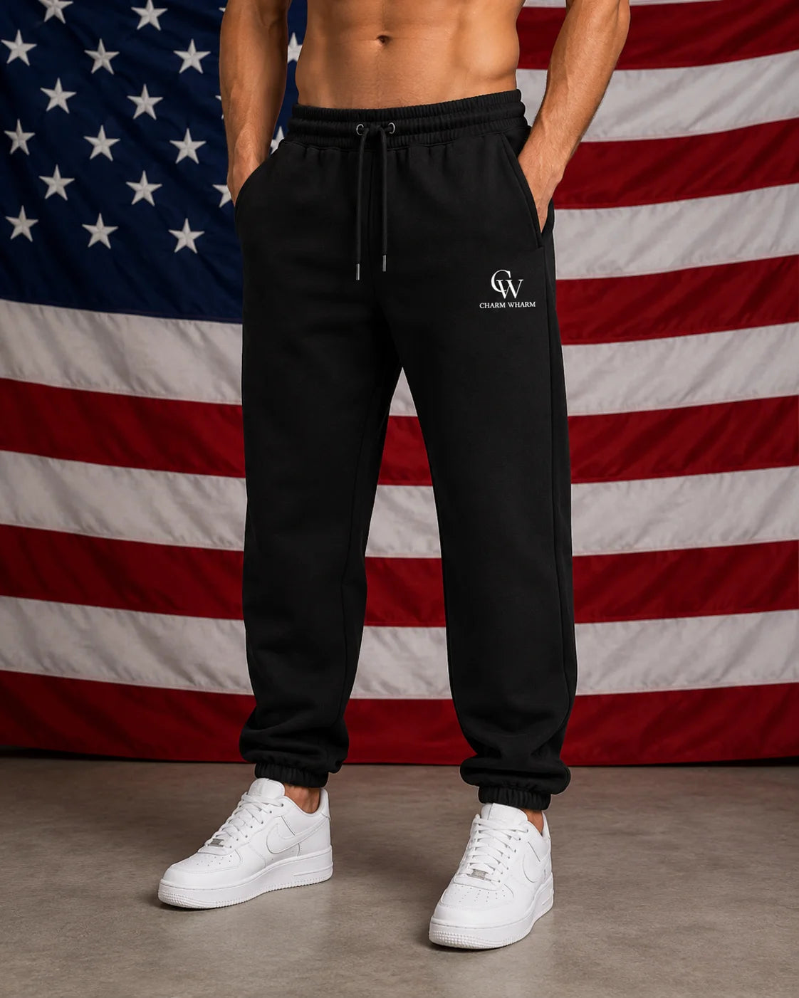 A man wearing black sweatpants with a white embroidered logo on the left thigh, standing in front of an American flag backdrop. The sweatpants have a drawstring waist and elastic cuffs, paired with white sneakers.