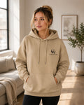 A woman wearing a beige pullover hoodie with a black embroidered 'CW' logo and 'CLOTHINGMADE' text on the left chest, standing in a bright, modern living room.