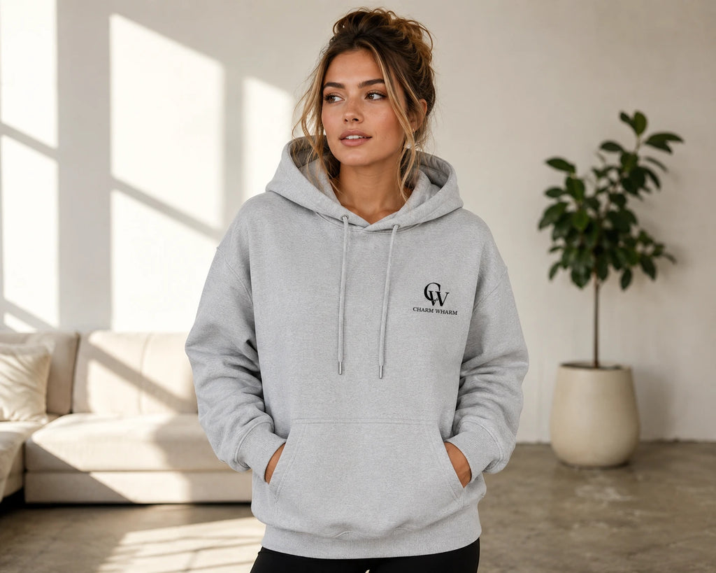 A woman wearing a light gray pullover hoodie with a black 'CW' monogram logo on the left chest, standing in a bright, modern living room with a sofa and a potted plant in the background.