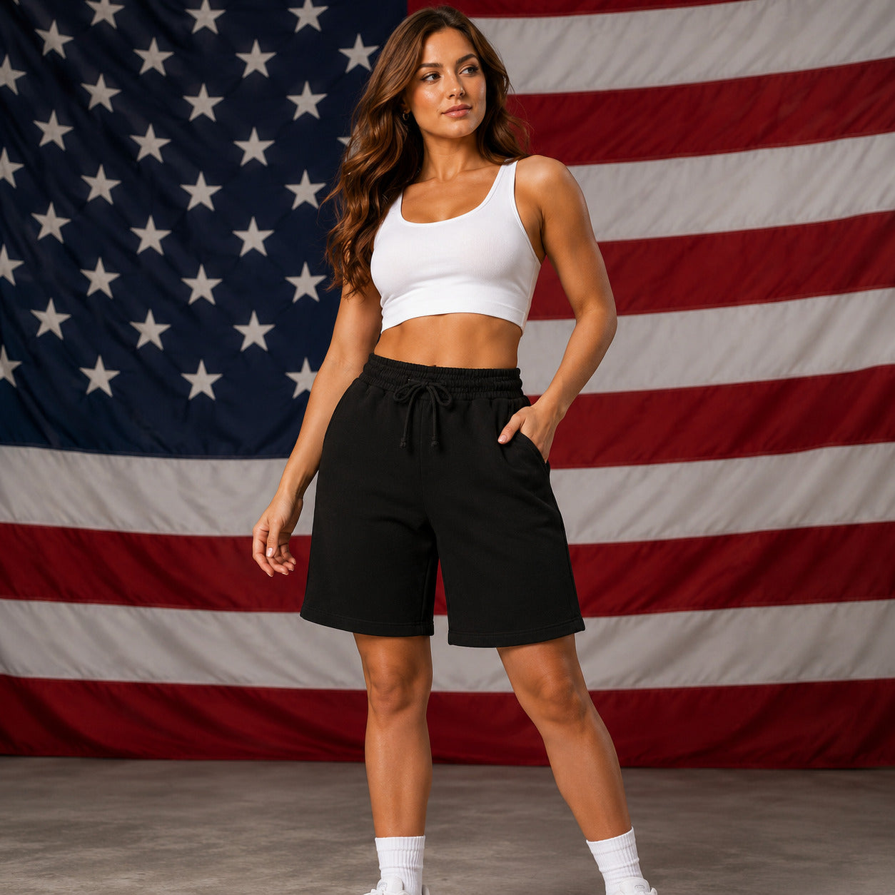 A woman modeling black drawstring shorts and a white crop top, standing in front of a large American flag backdrop.